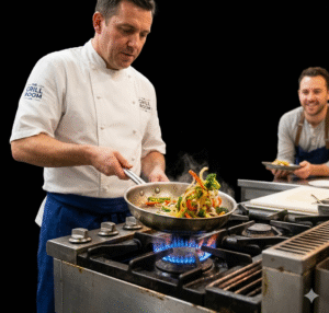 A professional photo of a chef with a focused expression, wearing a "THE GRILL ROOM" jacket, actively sautéing vegetables in a stainless steel skillet over a powerful, bright blue gas flame on a commercial kitchen range. His focus is on the pan, where steam is rising from the cooking food. In the right background, a smiling colleague looks on, holding a plate, providing a sense of a busy, successful SME environment. The blue flame from the range is prominent, highlighting the essential role of gas in this hospitality business.